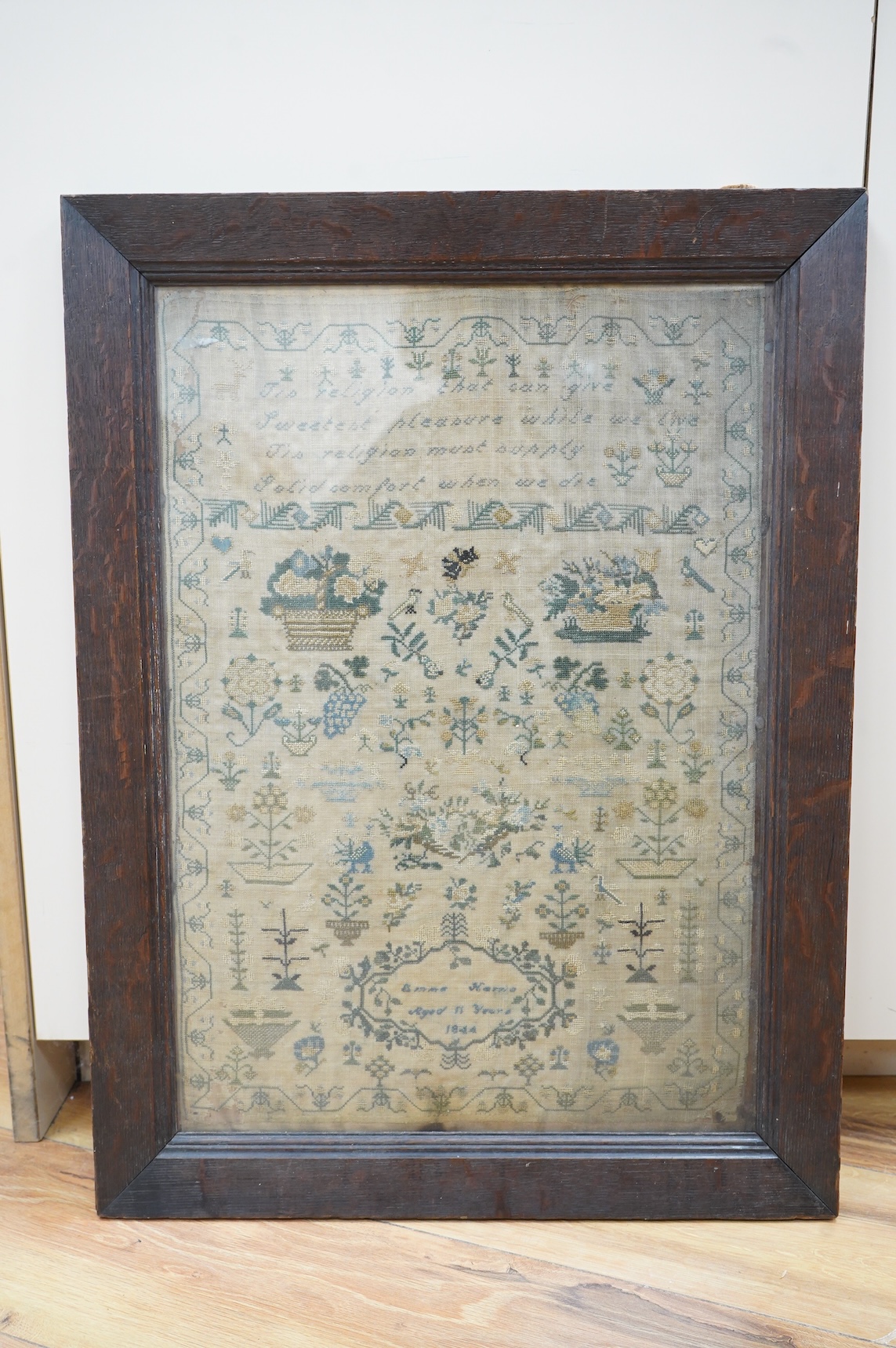 A framed 19th century sampler by Emma Harris, aged 11years, dated 1844. Worked with a verse and large baskets of flowers, with many spot motifs of flowers and birds amongst stylistic trees, worked within a vining border,